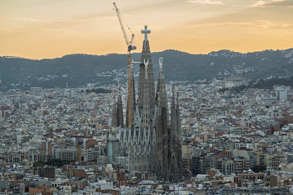 An aerial view of an ornate cathedral with tall spires, set against a dense urban cityscape at sunset.