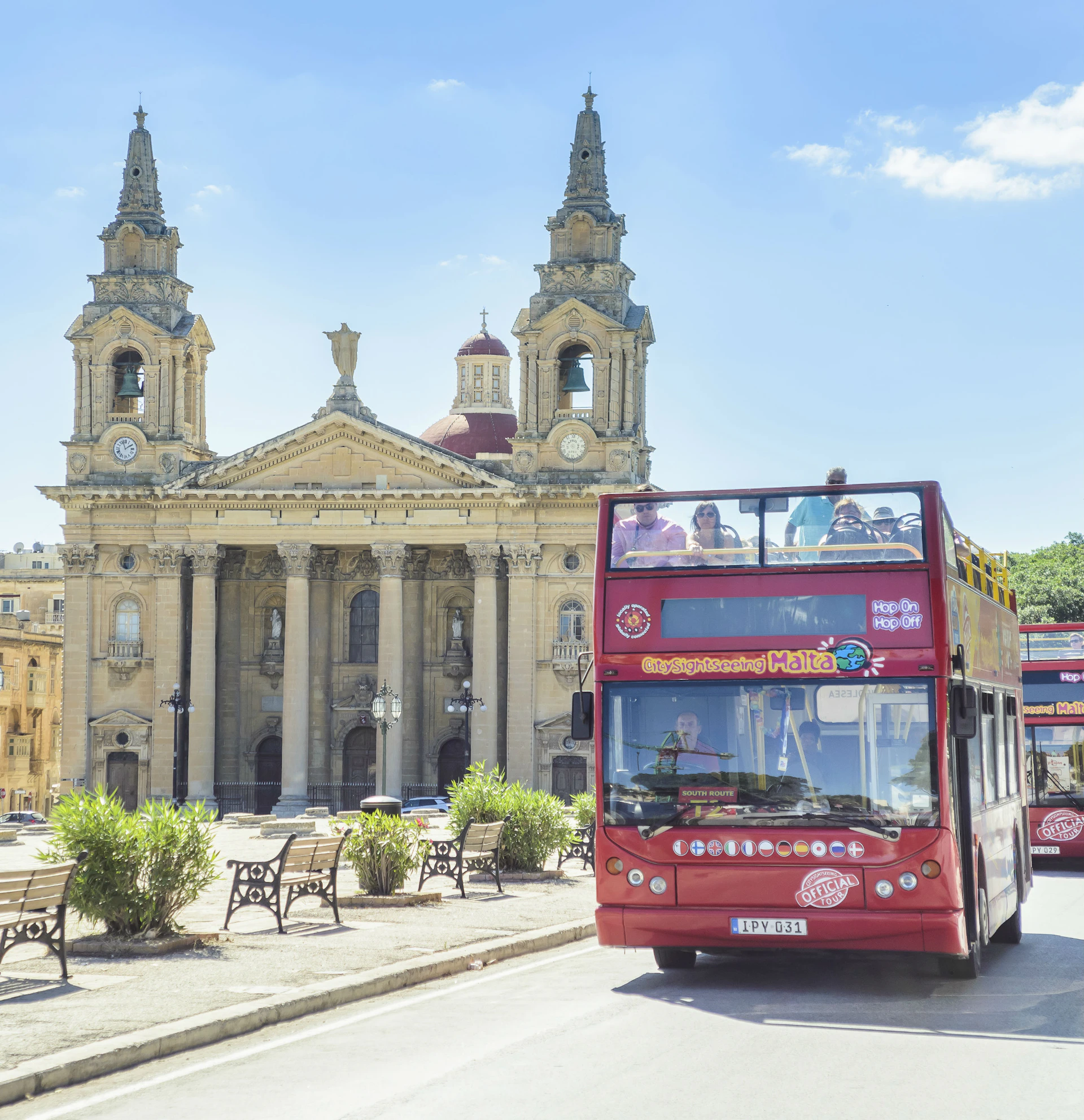 Red double-decker tour bus in front of a historic church with twin towers and benches on a sunny day.