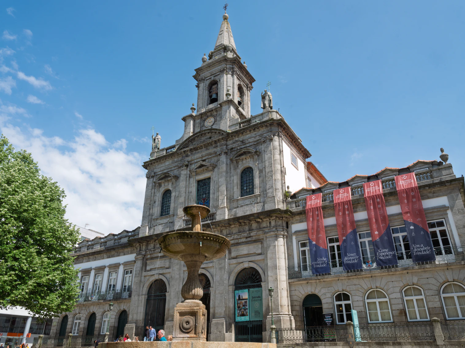 Historic stone building with a tall spire, central fountain in front, red banners hanging on both sides, and a blue sky above.