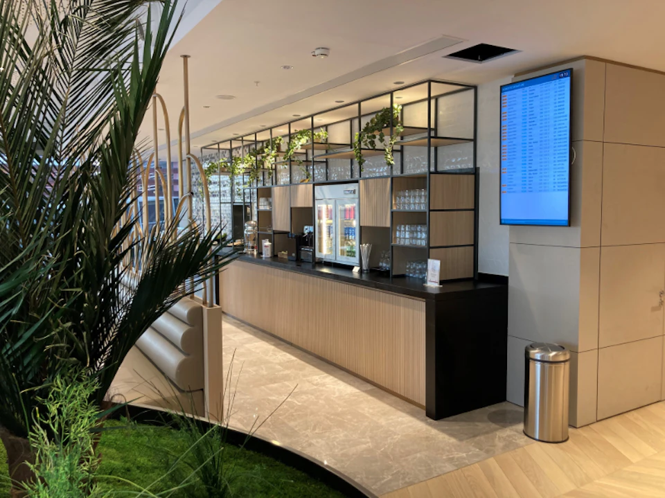 Modern coffee bar with wooden accents, hanging plants, and glass shelves. A digital display and trash can are on the right side.