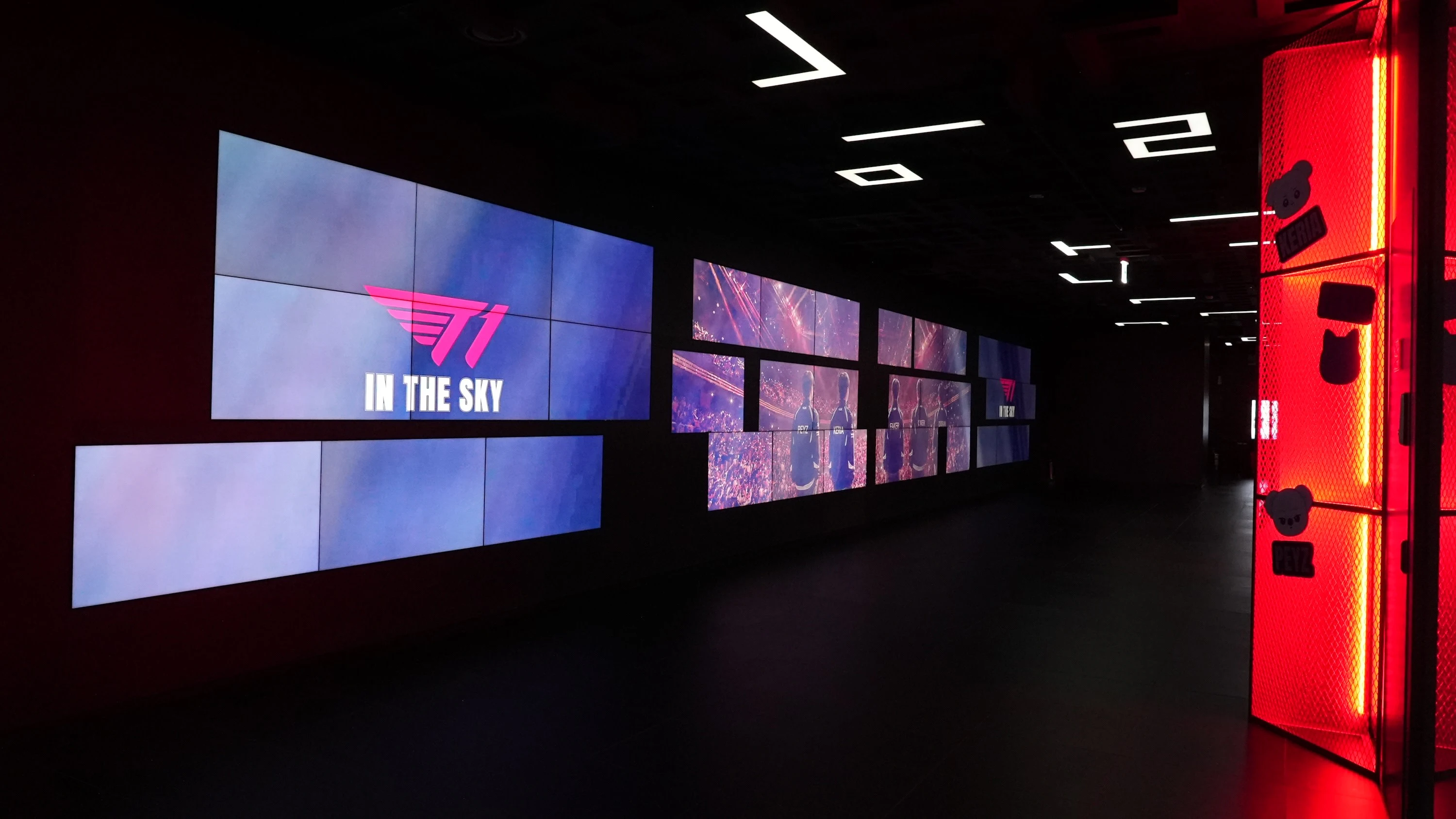 A wall of screens displays visuals and text, including "T1 IN THE SKY," in a dimly lit room.