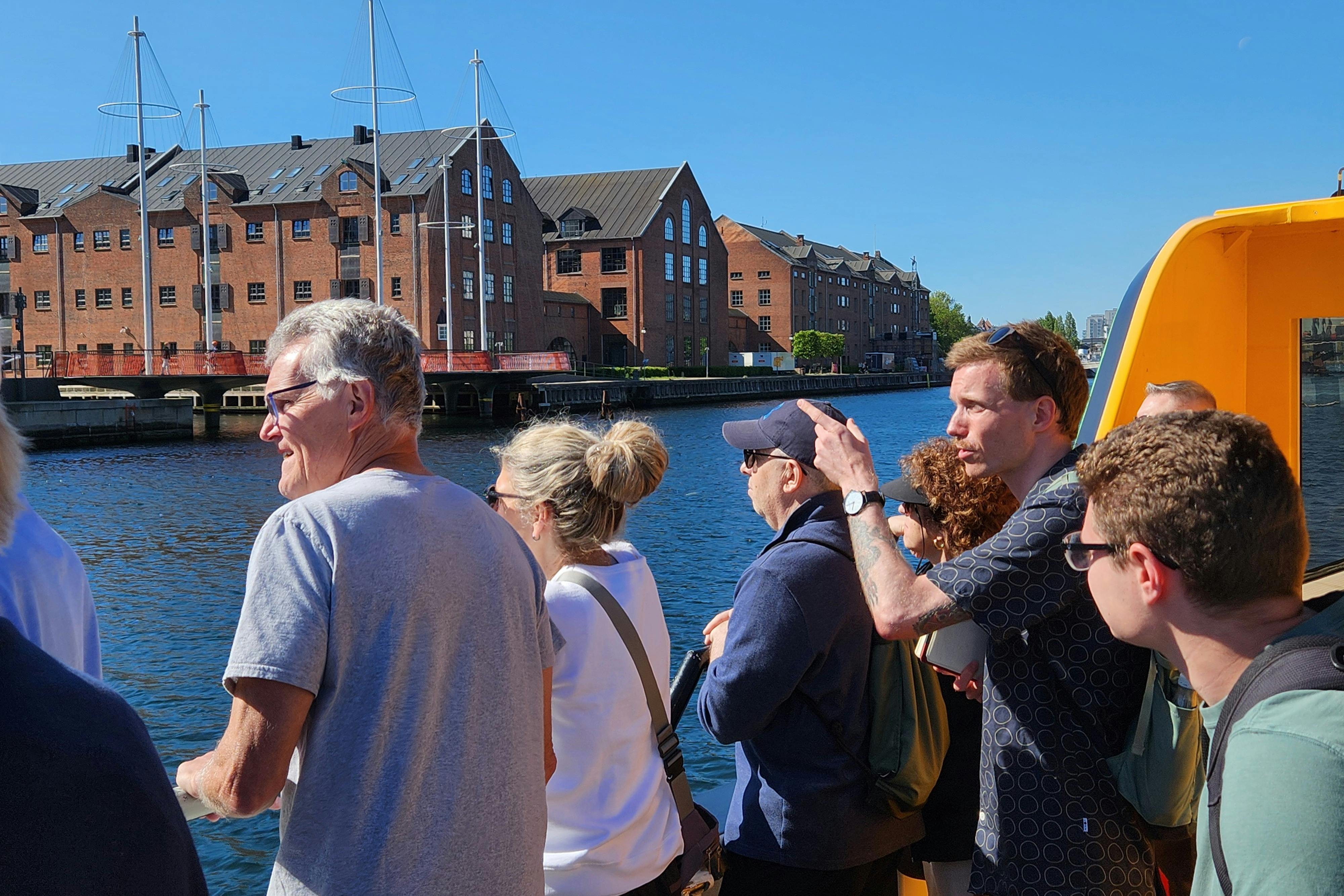 A group of people, some facing forward, on a boat near a water canal, with brick buildings and blue sky in the background.