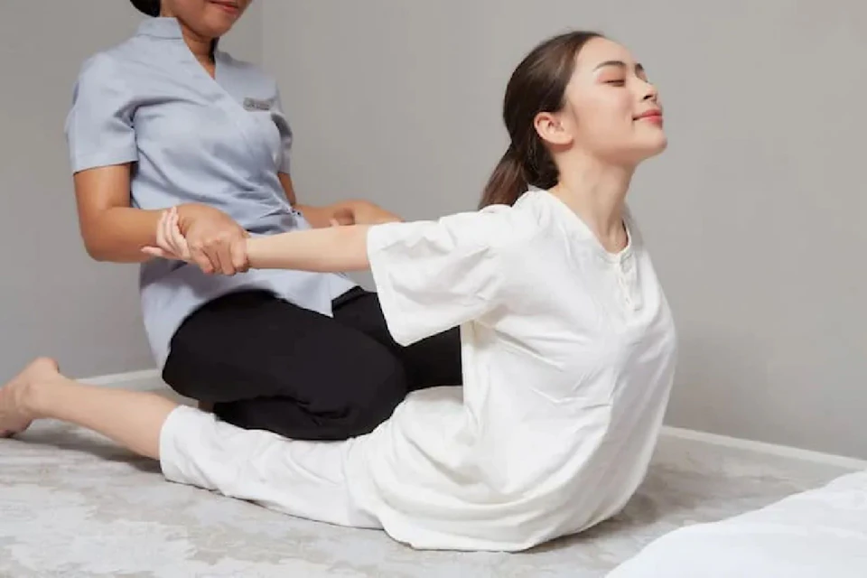 A person in a white outfit stretches their back on a mat while assisted by another person in a seated position.