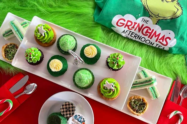 A festive dessert spread featuring green cupcakes, sandwiches, and a 'Grinch Afternoon' branded cloth on a green surface.
