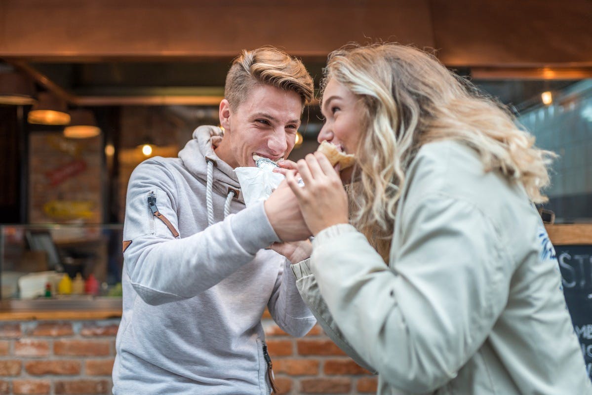 Δύο άνθρωποι μοιράζονται με χαρά φαγητό σε εξωτερικούς χώρους, ο ένας κρατώντας ένα τυλιγμένο αντικείμενο και ο άλλος τρώγοντας.