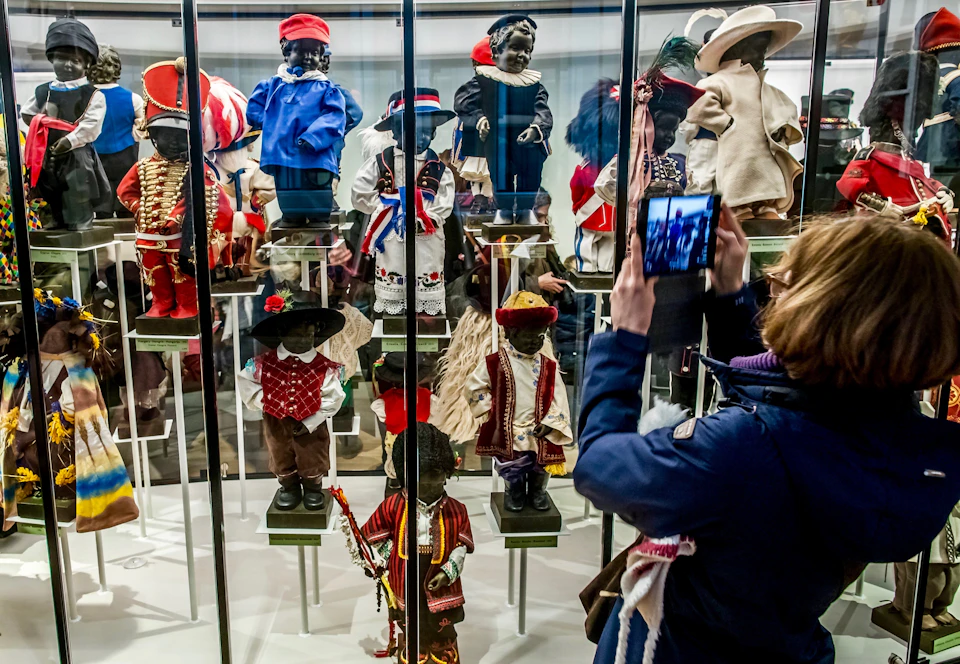 A person photographs a museum display of diverse, traditional dolls wearing various cultural outfits behind a glass case.
