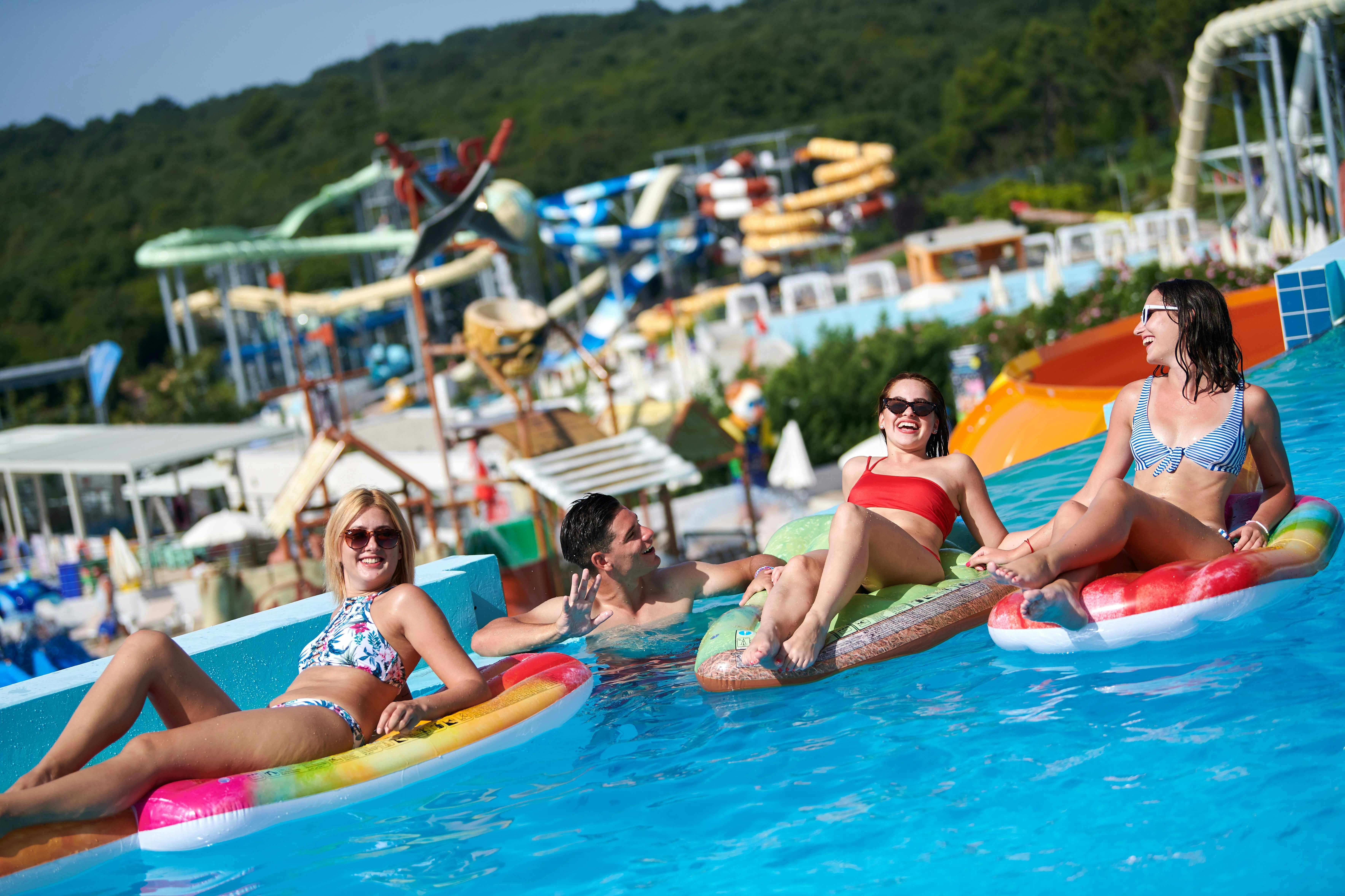 Vier lächelnde Menschen entspannen sich auf aufblasbaren Schwimmern in einem Pool in einem Wasserpark, mit bunten Rutschen und Grünpflanzen im Hintergrund.