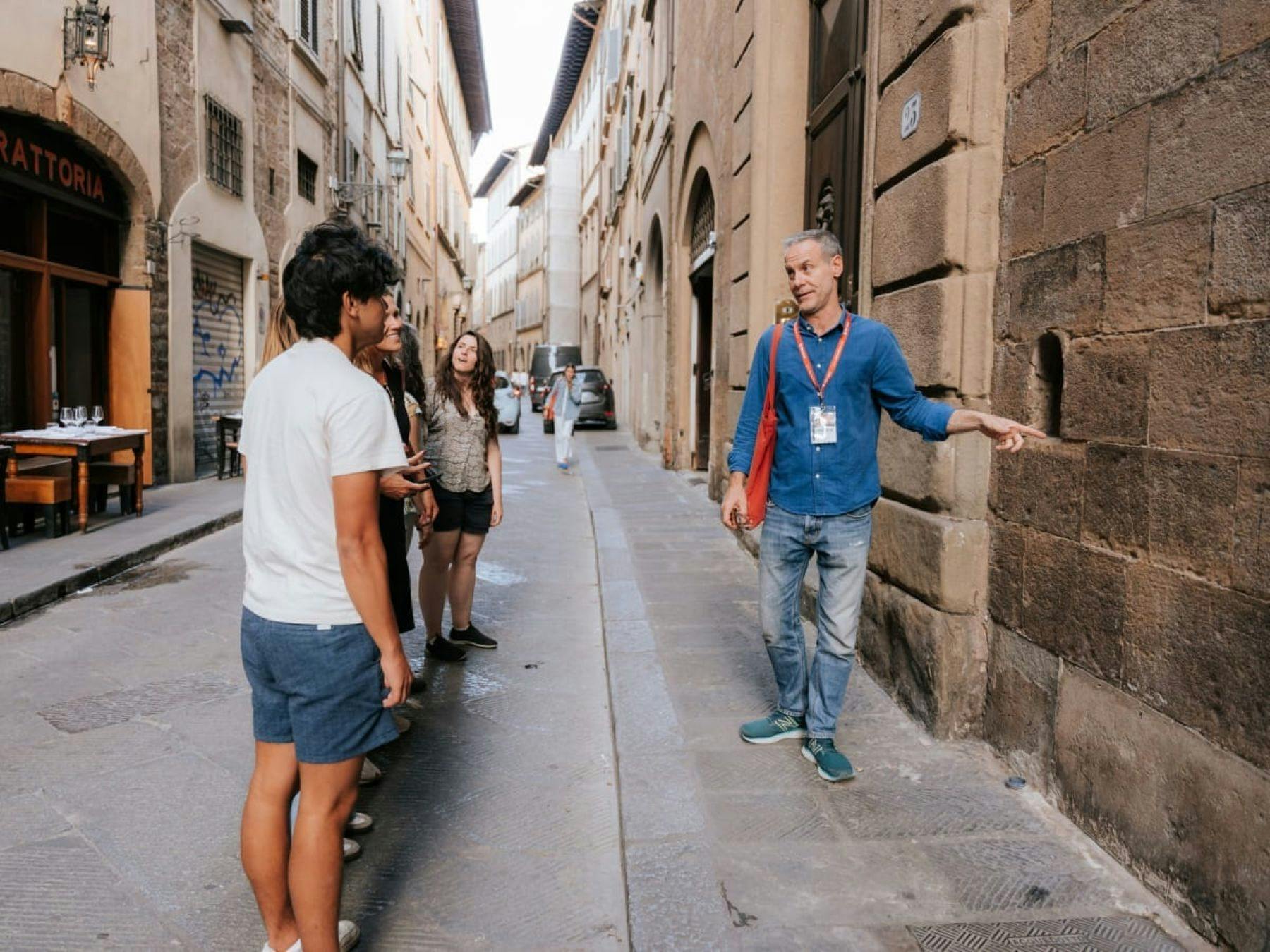 Una guida indica un dettaglio su un muro di pietra mentre un gruppo di persone ascolta in una stradina fiancheggiata da edifici storici.