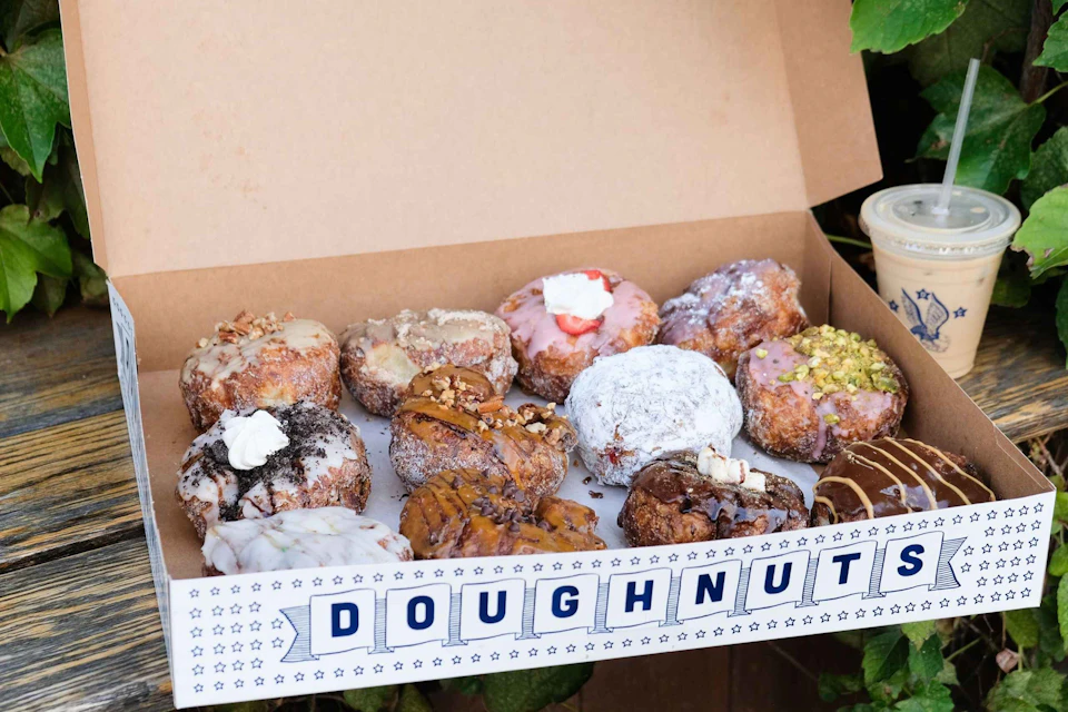 A box of assorted doughnuts, each topped with various toppings like cream, nuts, and powdered sugar, labeled "DOUGHNUT."