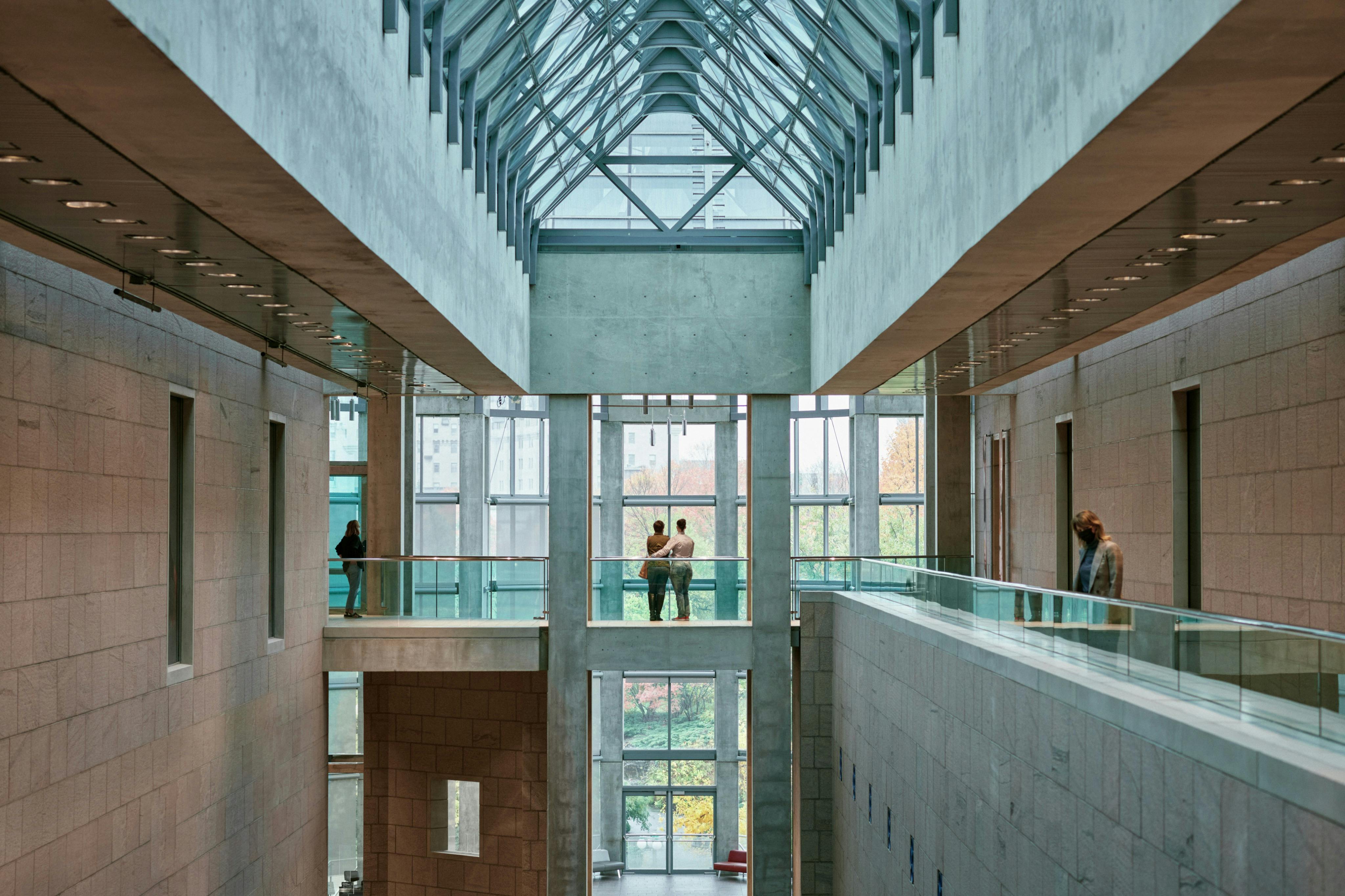 Glass-roofed atrium with several people standing by railings on different levels, overlooking a view of trees through large windows.