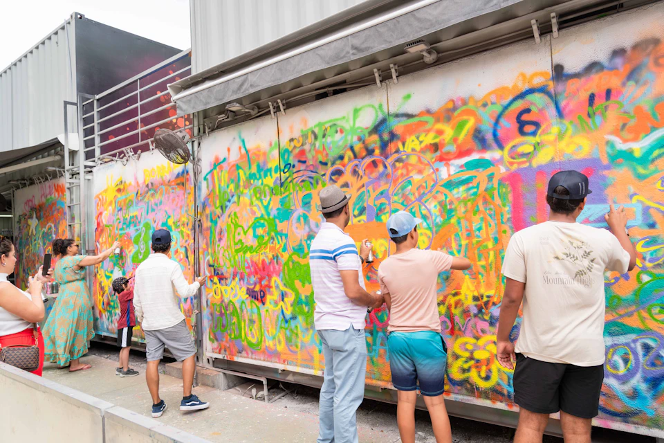 People painting colorful graffiti on an outdoor wall. Multiple layers of vibrant spray paint cover the entire surface.