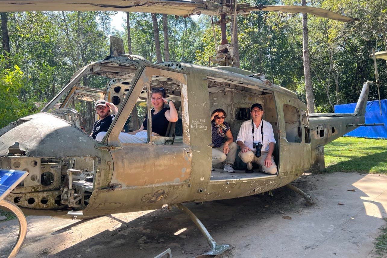 Four people sit inside an old, rusted helicopter fuselage in a wooded area, smiling for the camera.