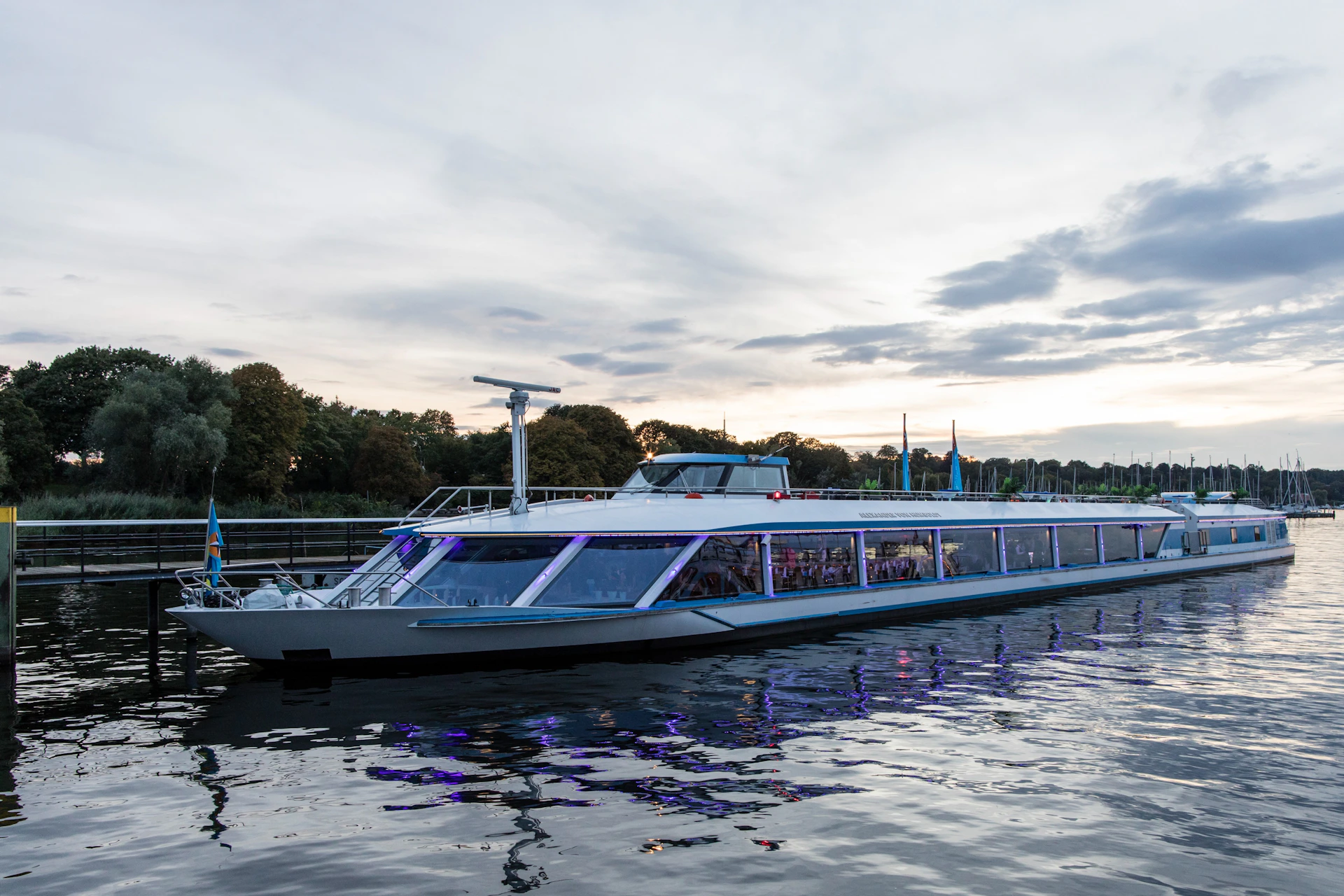 A modern glass-walled boat docked on calm water near a tree-lined shore at sunset, with reflections in the water.