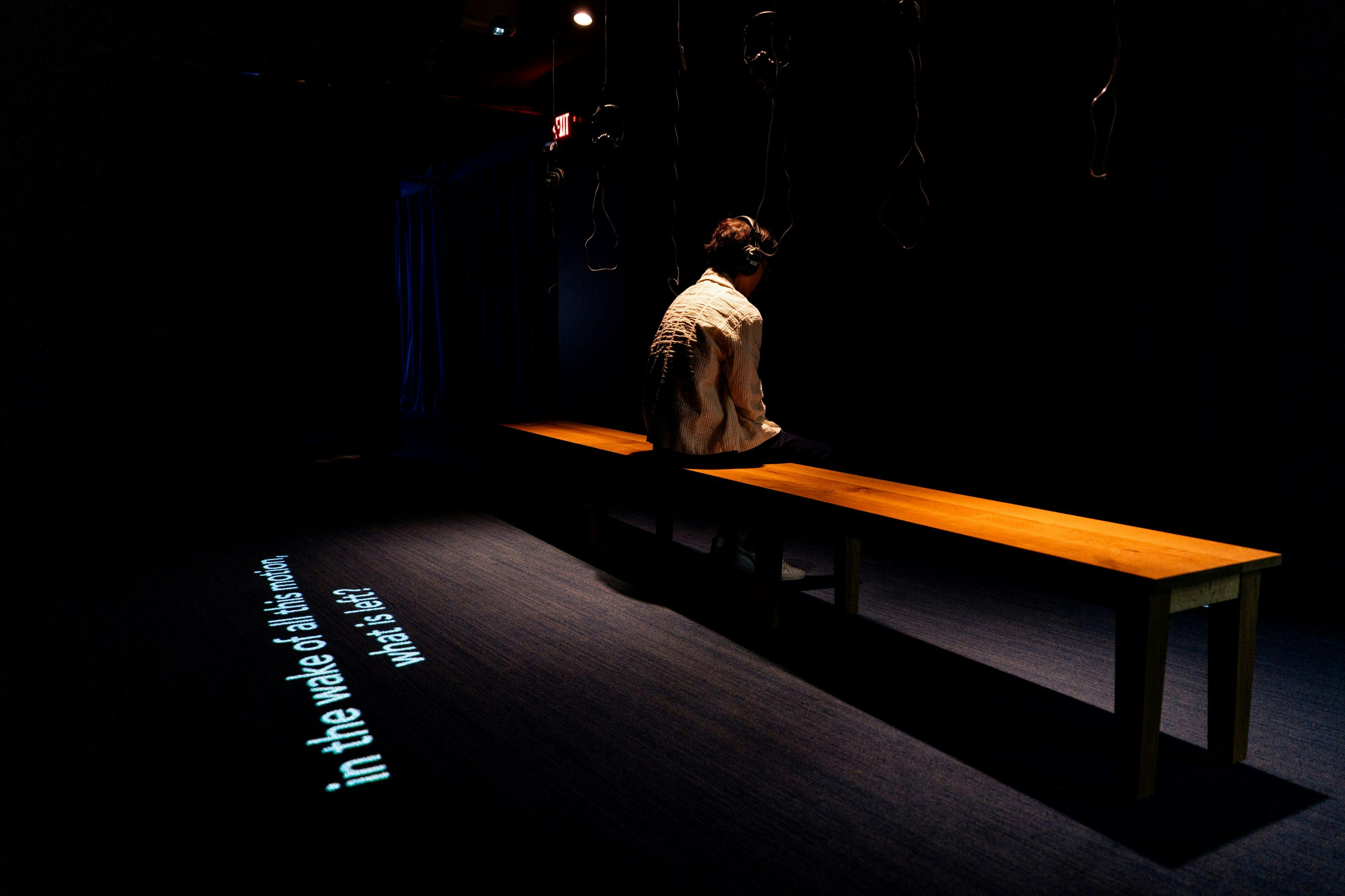 A person wearing headphones sits on a wooden bench in a dark room. Text is projected on the floor beside them.