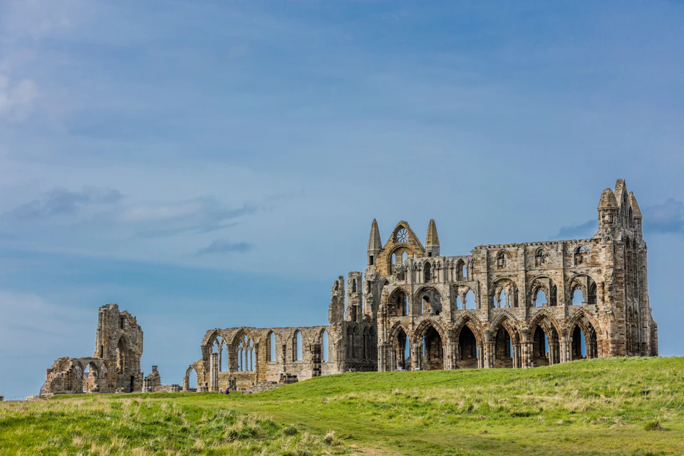 Ruins of an old stone abbey with tall arches and spires, situated on a grassy hill under a blue sky.