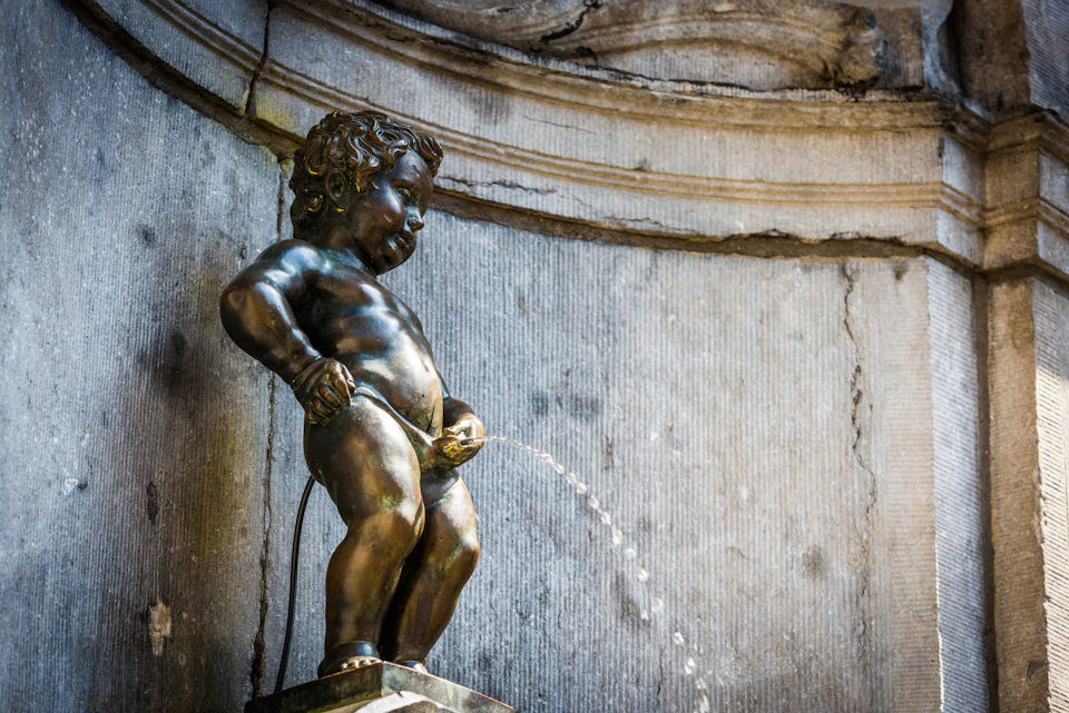 Bronze statue of a boy urinating, serving as a water fountain against a stone wall backdrop.
