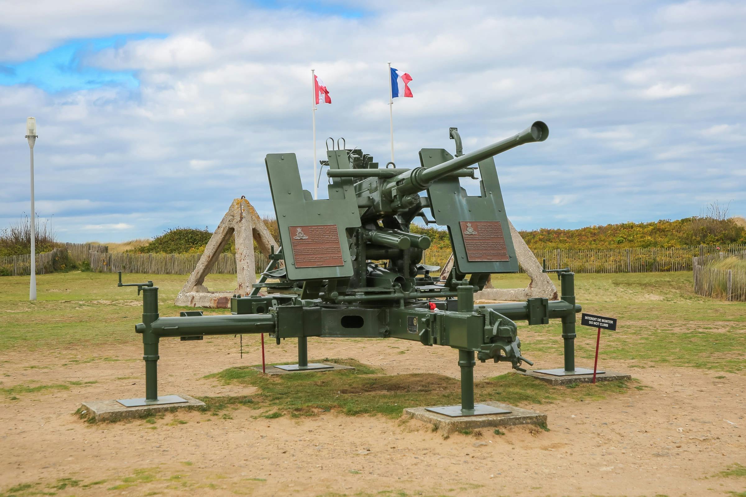 Ένα μεγάλο αντιαεροπορικό όπλο εμφανίζεται σε εξωτερικούς χώρους με σημαίες και ξύλινες κατασκευές στο παρασκήνιο κάτω από έναν μερικώς συννεφιασμένο ουρανό.