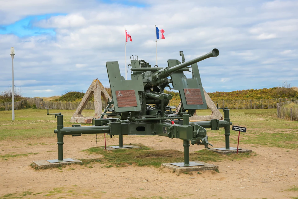 A large anti-aircraft gun is displayed outdoors with flags and wooden structures in the background under a partly cloudy sky.