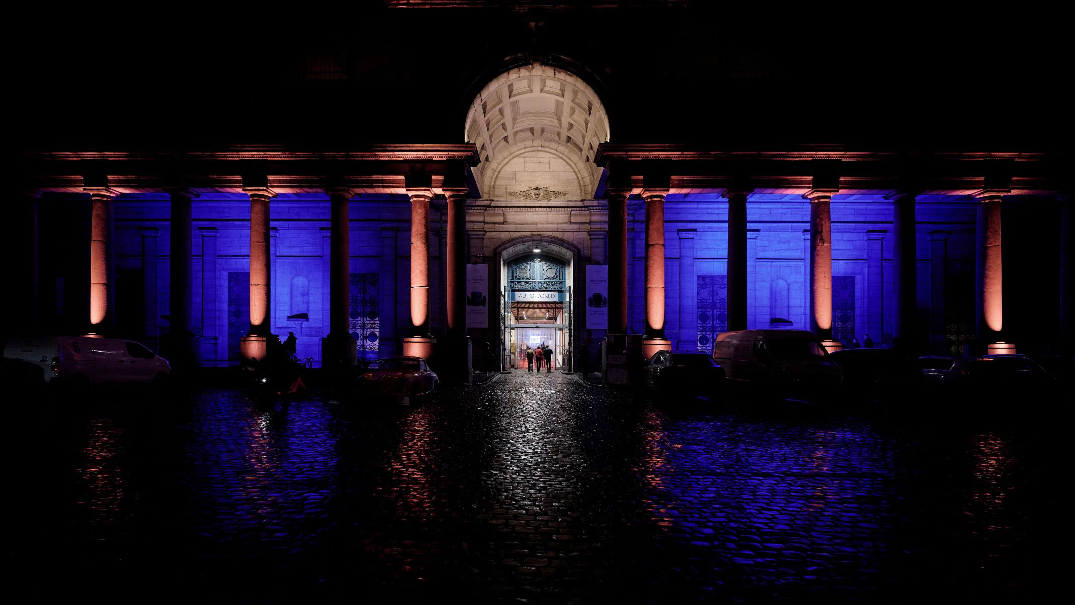 An illuminated building entrance at night with blue and orange lights, columns, and a cobblestone path. People enter in the distance.