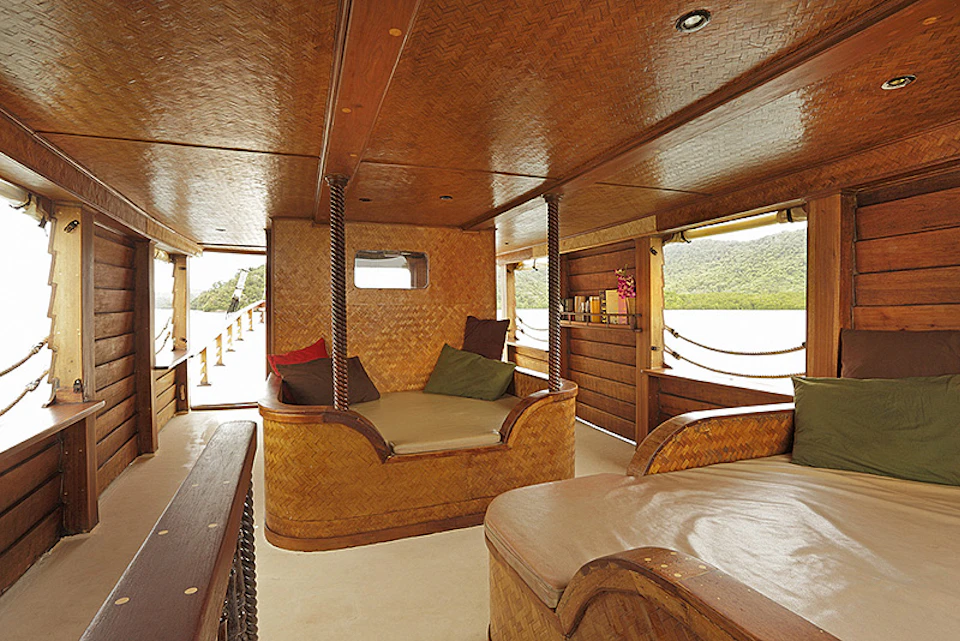 Wood-paneled boat interior with cushioned seating, pillows, and large windows offering a view of water and distant hills.