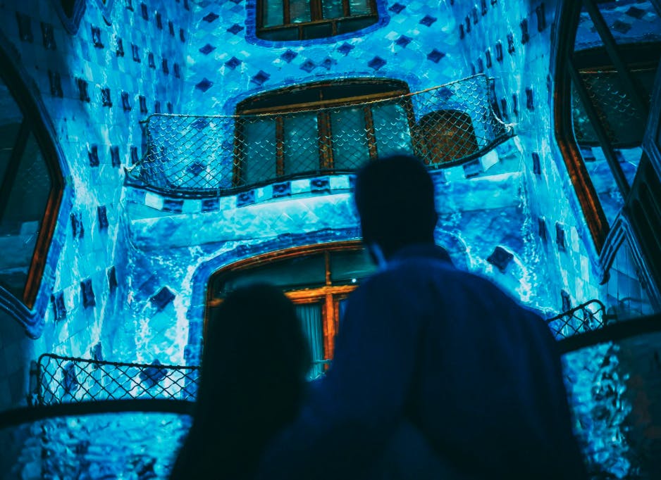 Two people viewing an illuminated blue-tiled building with intricate balconies and windows at night.