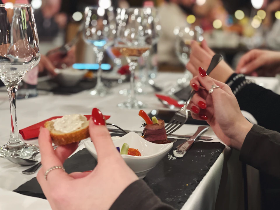 Close-up of hands with red nails holding a bread slice and fork, dining at a table set with wine glasses and small plates of food.