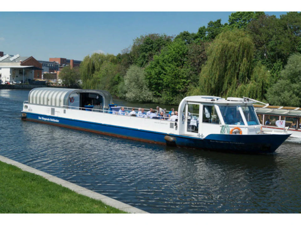 A white and blue canal boat with passengers travels along a tree-lined waterway on a sunny day.