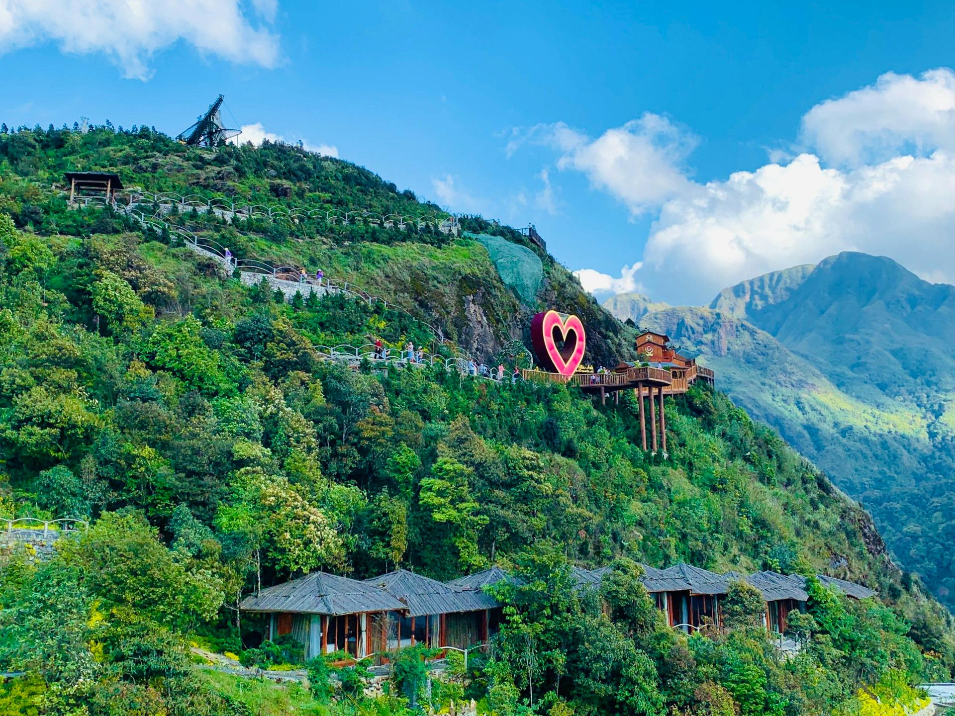 A scenic hillside with a heart-shaped decoration, traditional-style roofs below, and a wooden structure extending outward.