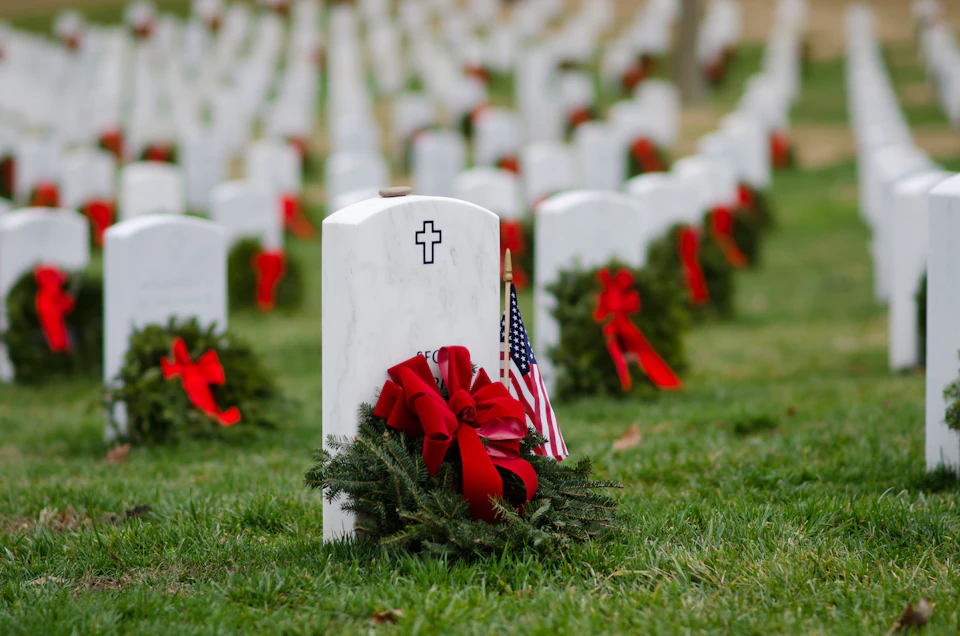 A white headstone in a cemetery, adorned with a wreath, red bow, and an American flag. Rows of similar graves with wreaths in the background.