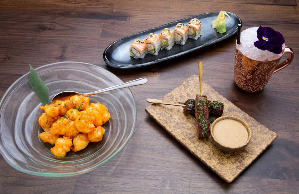 A dining setup featuring sushi rolls, shrimp dish, beef skewers with sauce, and a drink on a wooden surface.
