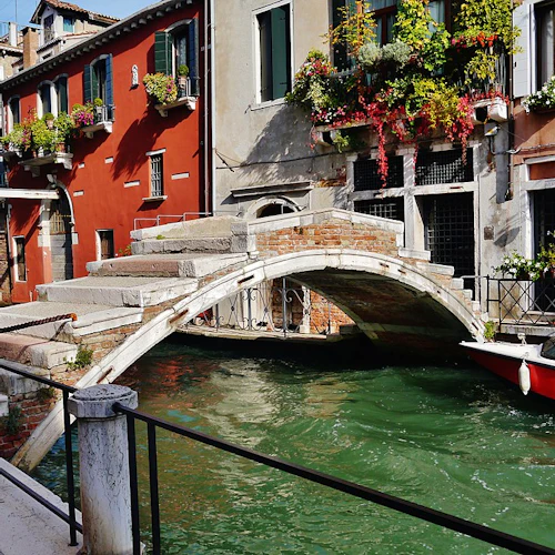 En stenbro över en kanal med färgglada byggnader och krukväxter; en röd båt är dockad till sidan.