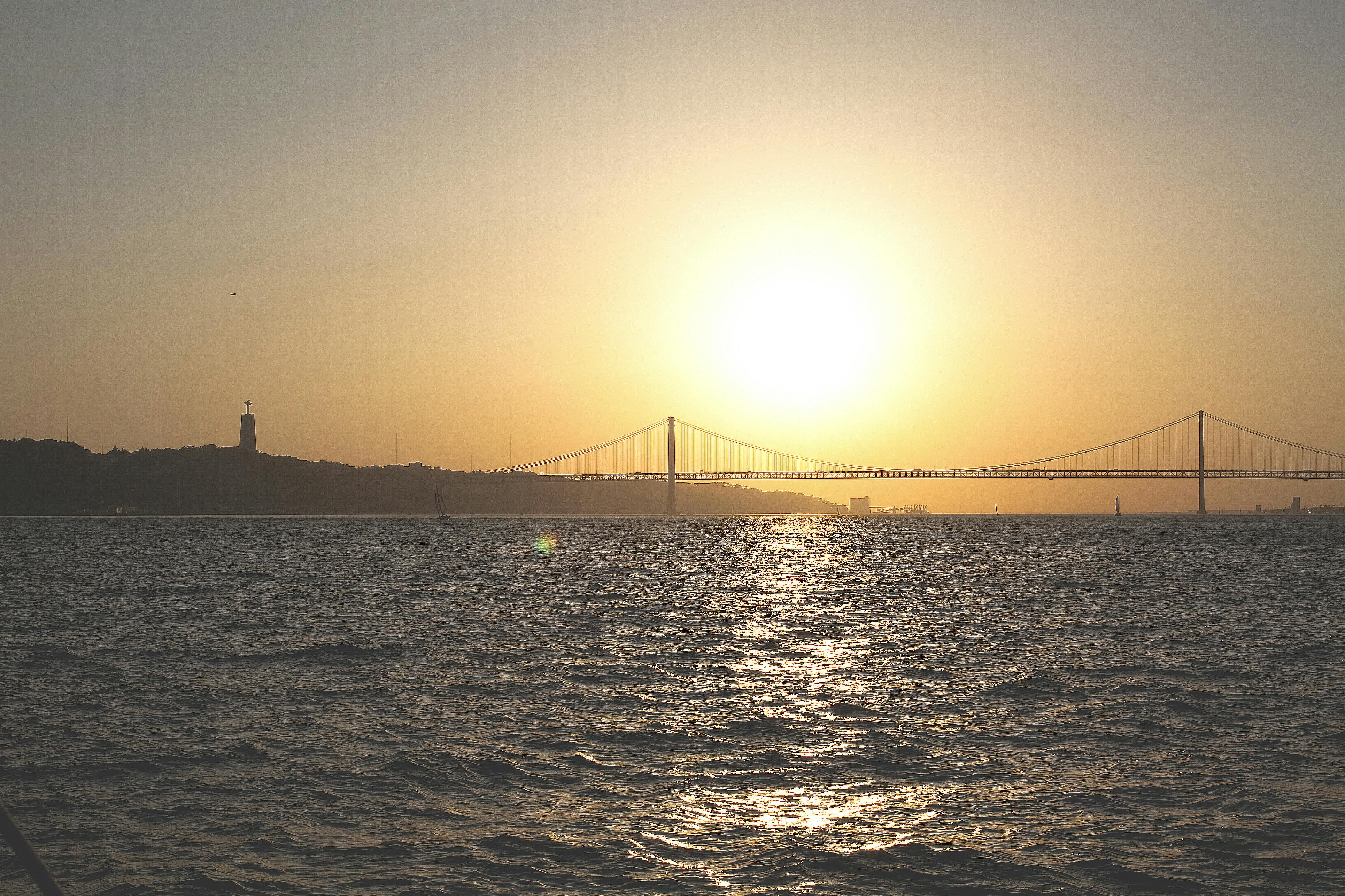 吊り橋は日没時に水の上に広がり、左側の地平線には像があり、頭上では太陽が明るく輝いています。