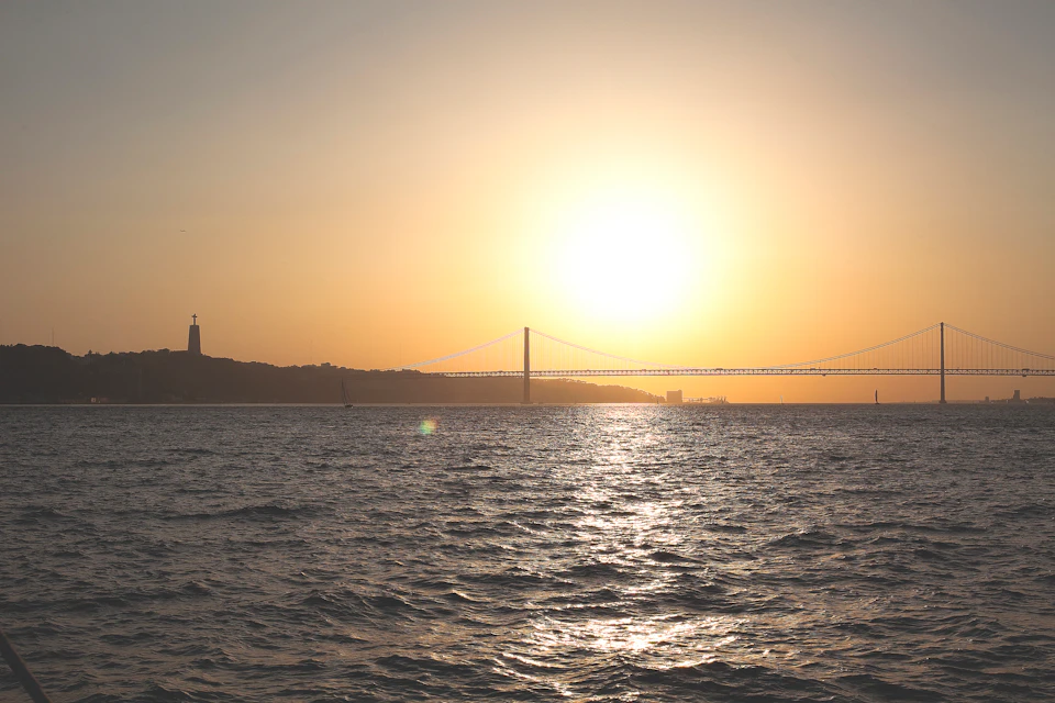 A suspension bridge extends over water at sunset, with a statue on the left horizon and the sun shining brightly overhead.