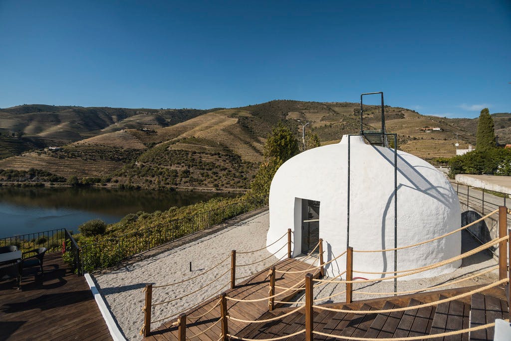 Een wit koepelvormig gebouw met een houten loopbrug tegen een achtergrond van heuvelachtig terrein en een helderblauwe lucht.