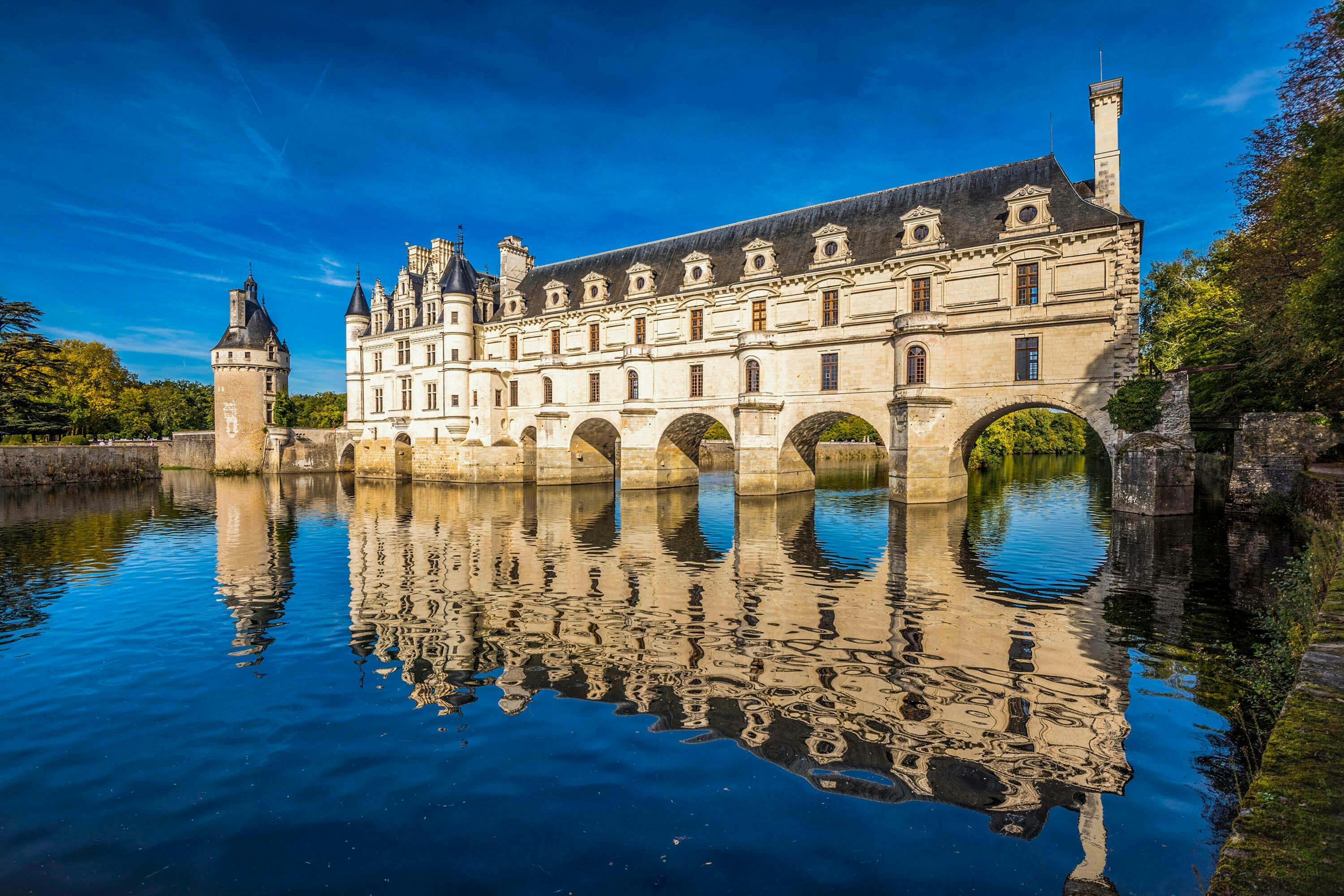 여러 개의 아치가 있는 돌다리가 잔잔한 강 위로 뻗어 있으며, 밝고 푸른 하늘 아래 커다랗고 화려한 성이 반사됩니다.