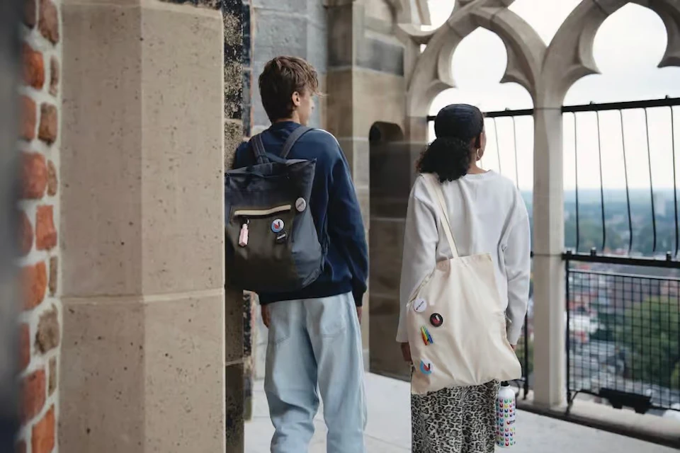 Two people with backpacks and tote bags stand on a building balcony with gothic-style architecture, looking outward.