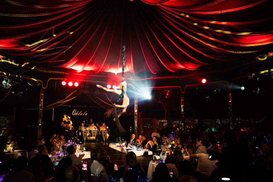 A performer on aerial silks under a red-lit tent, with an audience seated at tables watching the performance.