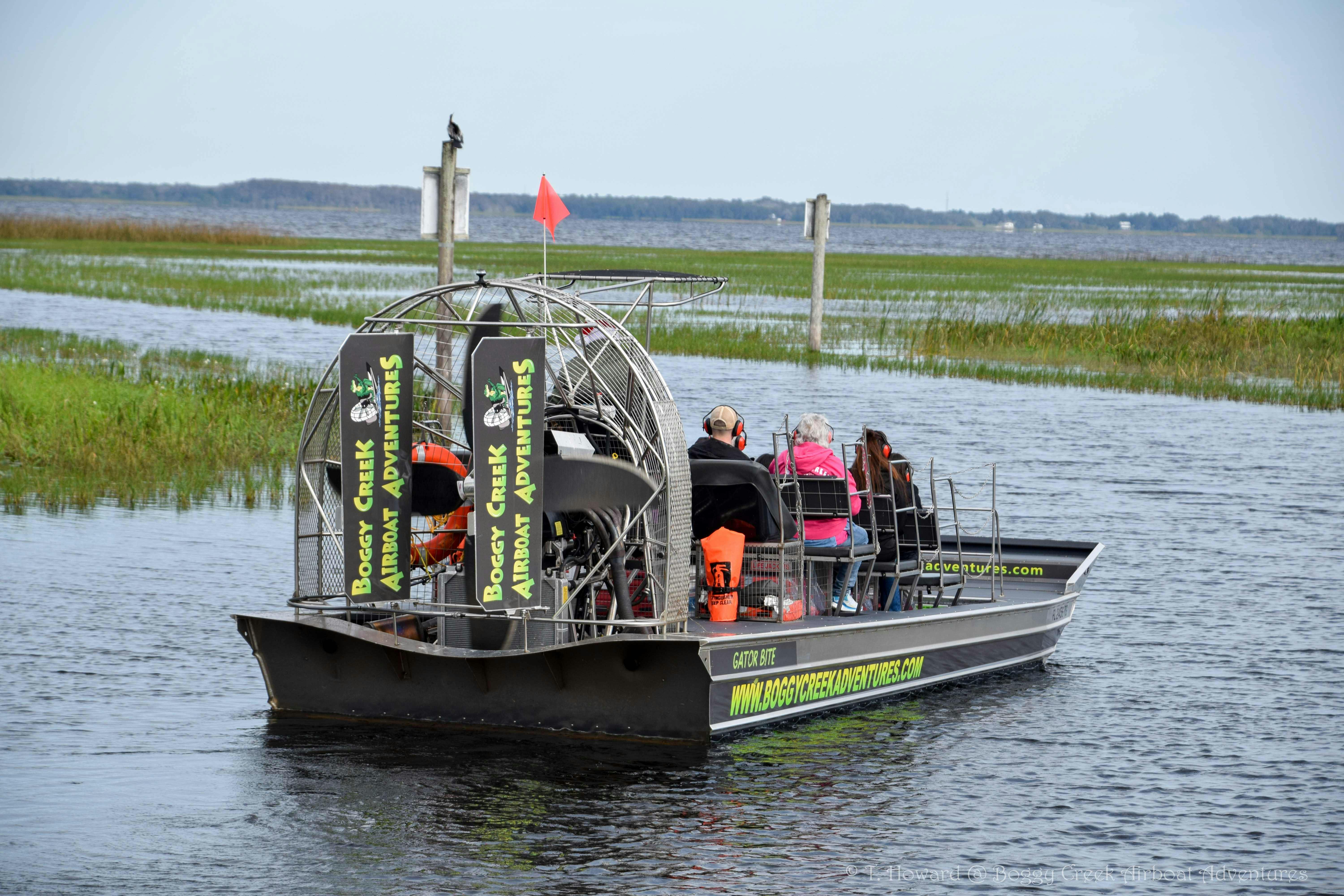 Un bateau pneumatique avec des passagers navigue dans un marais, avec des panneaux indiquant "Boggy Creek Airboat Adventures" (aventures en bateau pneumatique de Boggy Creek).