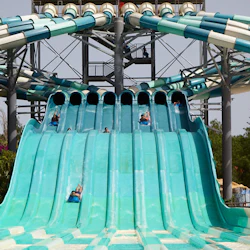 People sliding down multiple parallel water slides at a water park, with a tall structure in the background.
