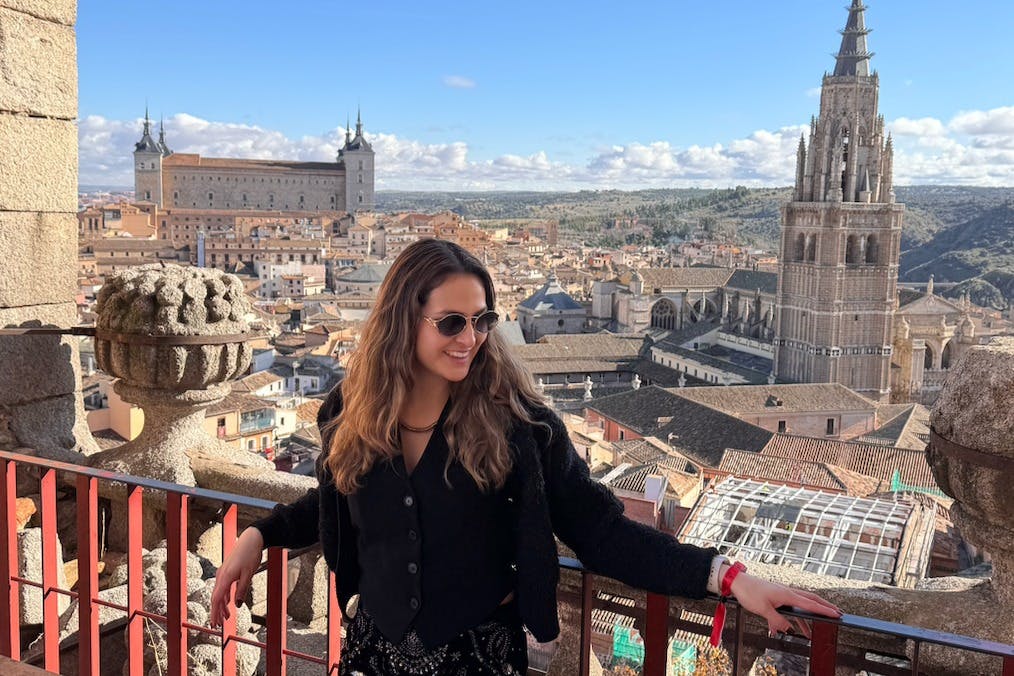 Une femme debout sur un balcon surplombant un paysage urbain pittoresque avec des bâtiments historiques et un ciel bleu clair.