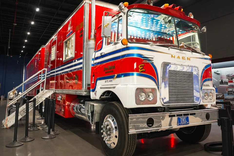 A bright red and white Mack truck with a custom design, parked indoors, featuring blue accents and stairs on the side.