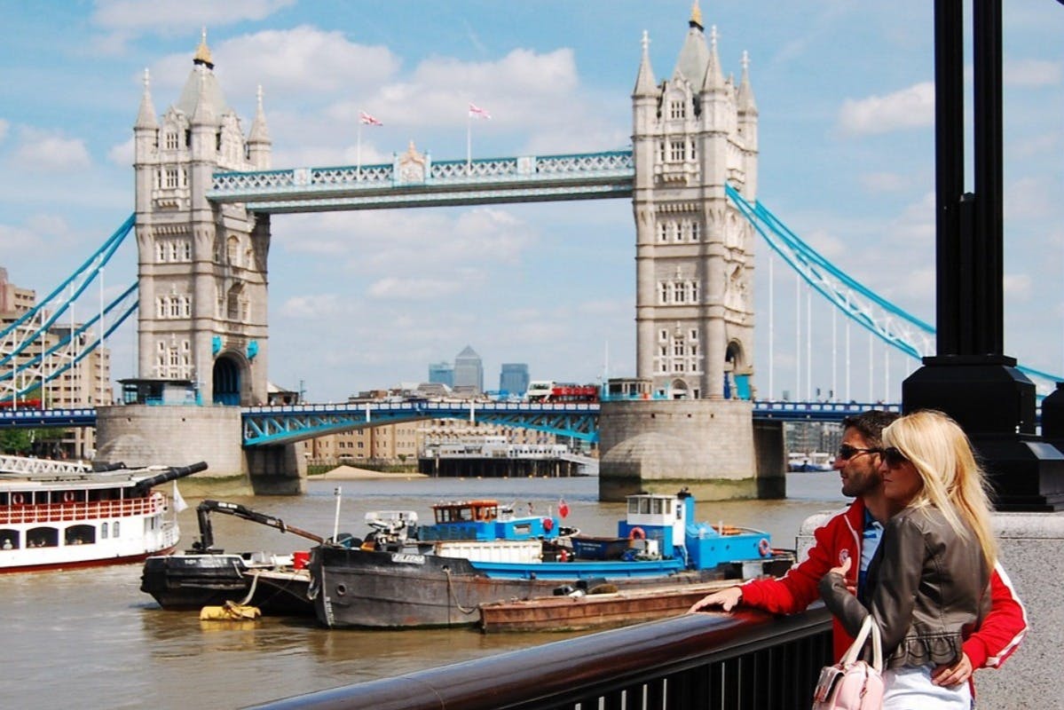 A couple stands by the Thames River, with Tower Bridge in the background on a partly cloudy day.