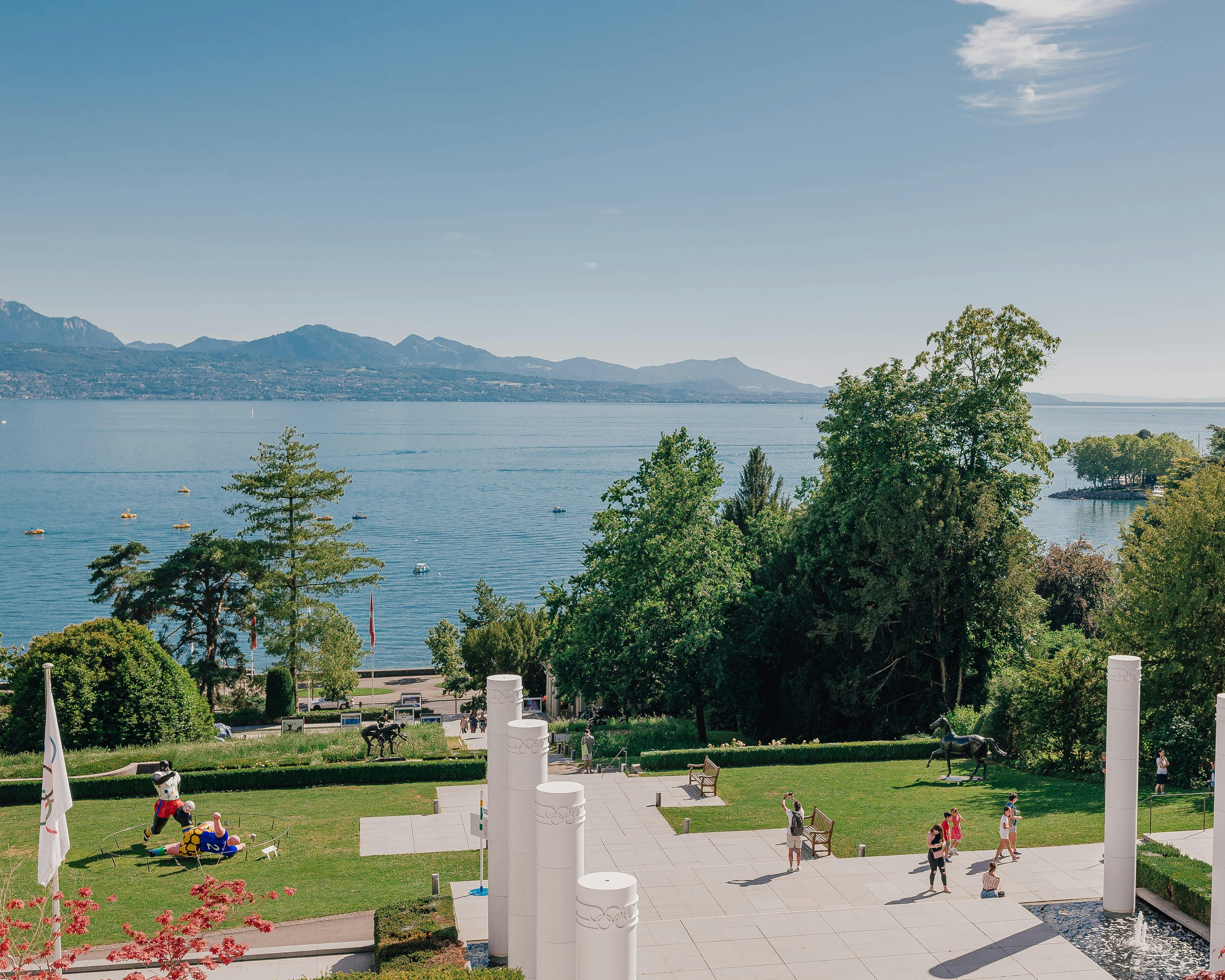 Lidé, kteří se procházejí a relaxují u jezera se stromy pokrytými kopci v pozadí pod jasnou modrou oblohou.