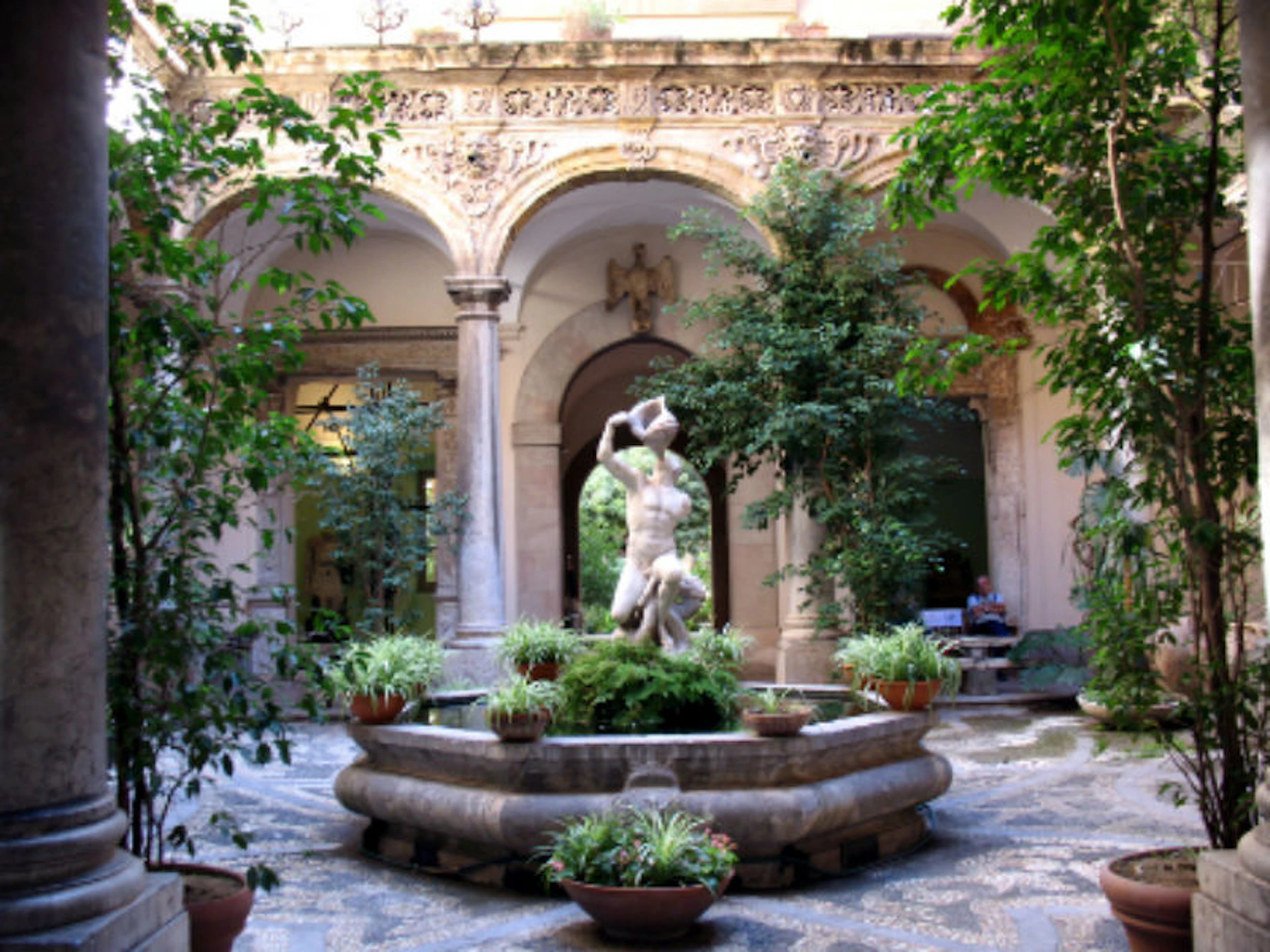 A courtyard with a central statue fountain, surrounded by greenery, columns, and arched walls with ornate designs.