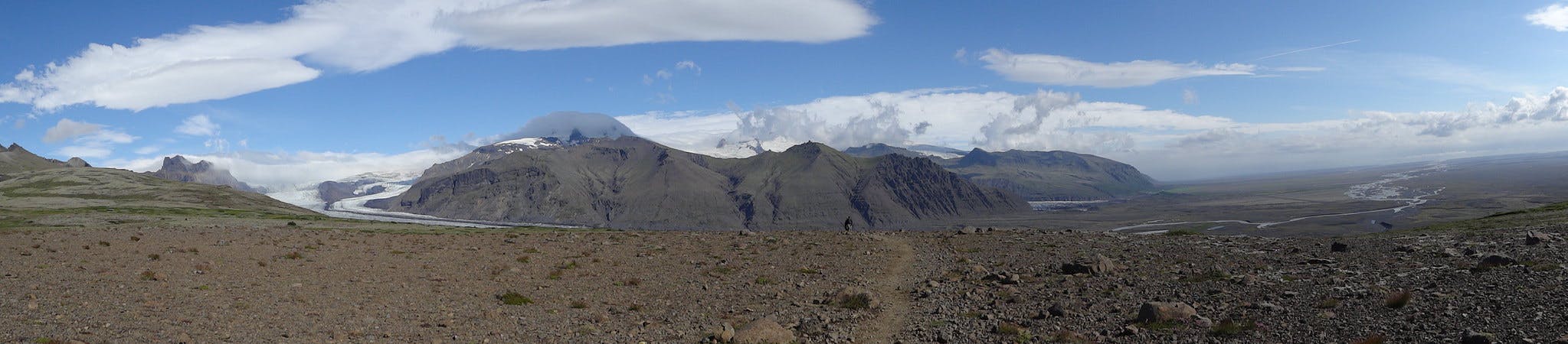 Panoramatický výhled na skalnatou krajinu se vzdálenými ledovci a horami pod modrou oblohou s rozptýlenými mraky.