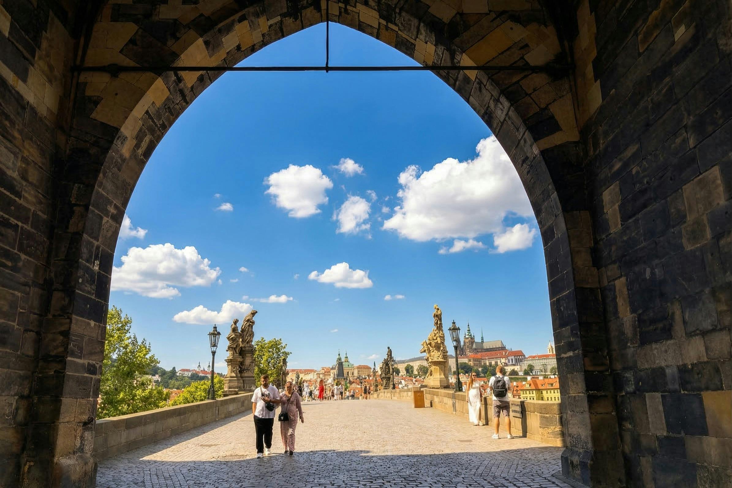 A stone archway frames a sunny view of a bridge with statues, people walking, and a cityscape in the background.