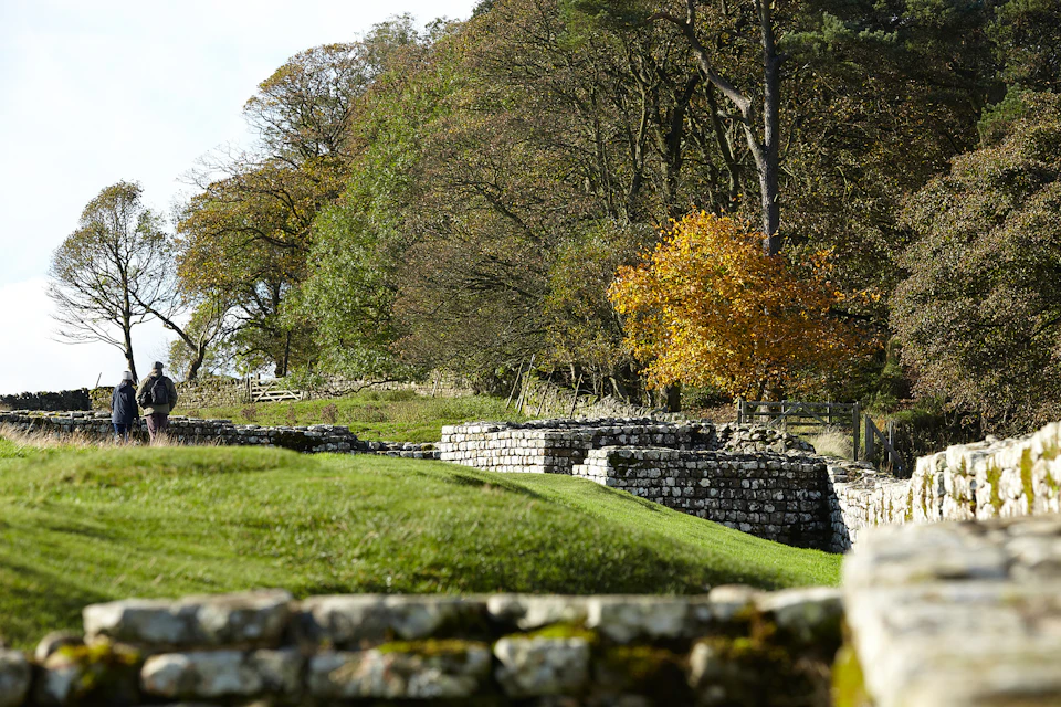 Ancient stone walls surrounded by green grass and trees with autumn foliage, under a partly cloudy sky.