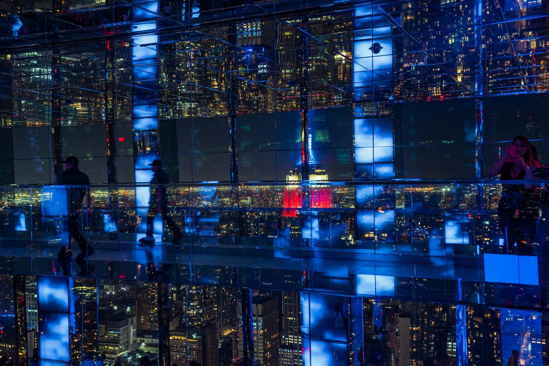 Une personne se promène dans une pièce à miroirs donnant sur un paysage urbain animé de nuit, avec une tour rouge brillamment éclairée visible.