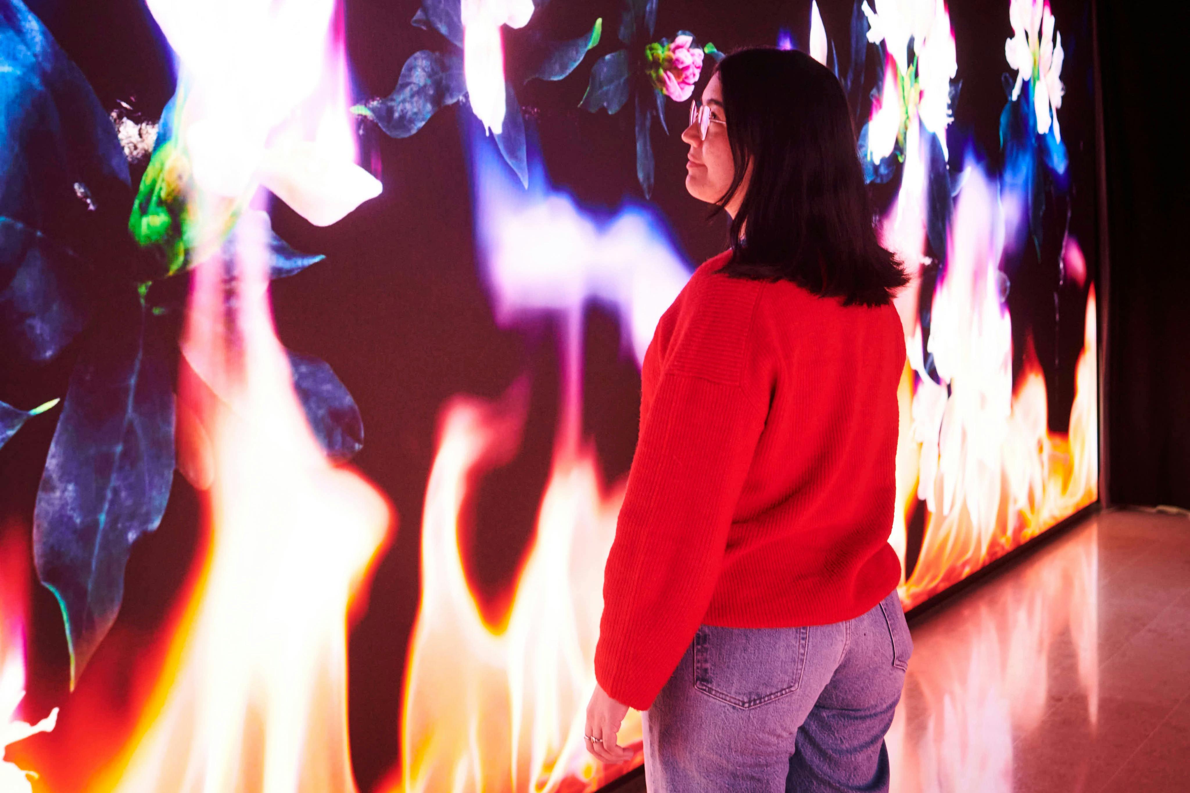 Kobieta w czerwonym swetrze i dżinsach stoi przed cyfrowym pokazem kolorowych płomieni i kwiatów.