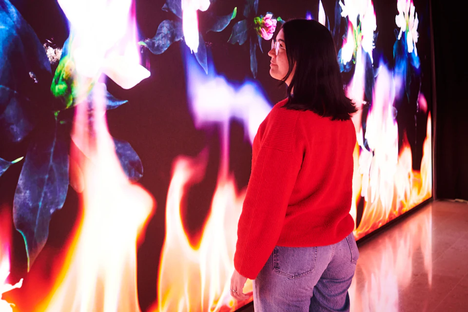 A woman in a red sweater and jeans stands facing a digital art display of colorful flames and flowers.